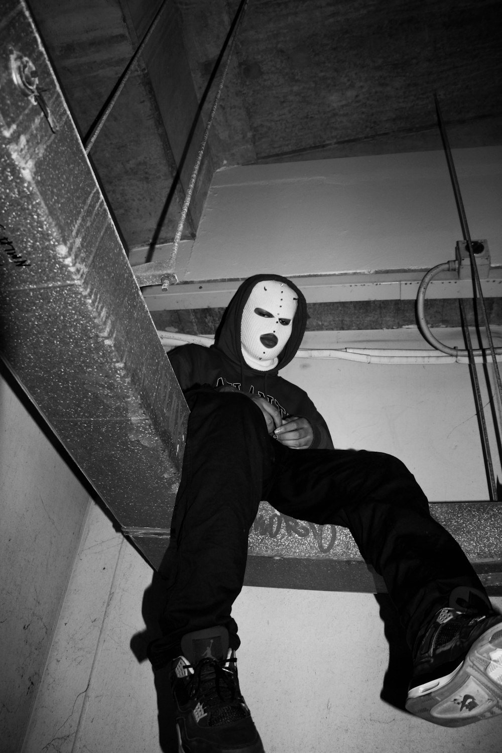 grayscale photo of man in black pants and white mask sitting on stairs