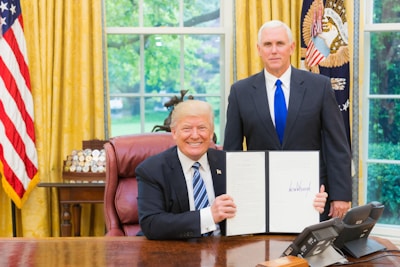 Two men in formal attire are in an office setting. One man is seated at a large wooden desk, holding up an open binder with a signature visible. The other man stands beside the desk. Behind them is a large window with a view of greenery, and an American flag drapes from the wall. The room features gold curtains and various decorative items.