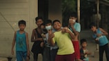 A group of children stand together outdoors, with many of them smiling and appearing playful. Some are wearing masks while others are casually dressed in various styles and colors of clothing. The setting appears to be a neighborhood with a building and trees visible in the background.