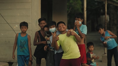 A group of children stand together outdoors, with many of them smiling and appearing playful. Some are wearing masks while others are casually dressed in various styles and colors of clothing. The setting appears to be a neighborhood with a building and trees visible in the background.