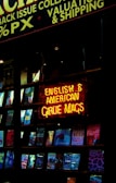 A shop window displaying a collection of magazines with a prominent neon sign in bright red and yellow. The sign reads 'English & American Girlie Mags.' Various magazine covers are visible, showcasing different themes and colorful designs.