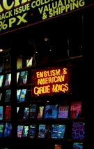 A shop window displaying a collection of magazines with a prominent neon sign in bright red and yellow. The sign reads 'English & American Girlie Mags.' Various magazine covers are visible, showcasing different themes and colorful designs.