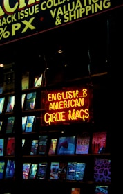 A shop window displaying a collection of magazines with a prominent neon sign in bright red and yellow. The sign reads 'English & American Girlie Mags.' Various magazine covers are visible, showcasing different themes and colorful designs.