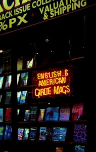 A shop window displaying a collection of magazines with a prominent neon sign in bright red and yellow. The sign reads 'English & American Girlie Mags.' Various magazine covers are visible, showcasing different themes and colorful designs.