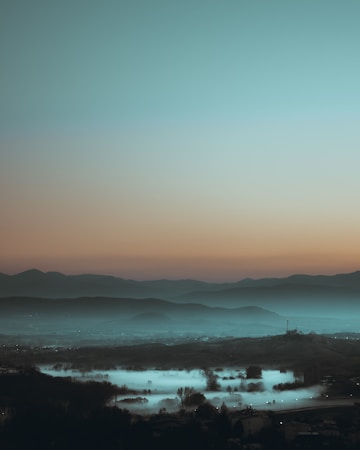 A serene landscape featuring mist-covered fields, silhouetted trees, and a range of mountains under a gradient sky transitioning from a soft orange to blue.