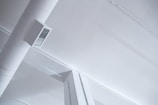 A modern, industrial-style ceiling with visible ductwork and air vents. The ceiling is painted white, and the duct is a cylindrical shape attached with a rectangular vent cover. The aesthetic features clean lines and minimalistic design.