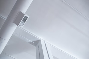 A modern, industrial-style ceiling with visible ductwork and air vents. The ceiling is painted white, and the duct is a cylindrical shape attached with a rectangular vent cover. The aesthetic features clean lines and minimalistic design.
