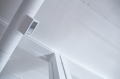 A modern, industrial-style ceiling with visible ductwork and air vents. The ceiling is painted white, and the duct is a cylindrical shape attached with a rectangular vent cover. The aesthetic features clean lines and minimalistic design.