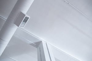 A modern, industrial-style ceiling with visible ductwork and air vents. The ceiling is painted white, and the duct is a cylindrical shape attached with a rectangular vent cover. The aesthetic features clean lines and minimalistic design.