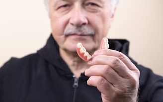 An older adult wearing a black jacket holds a set of dentures in their hand, with the focus on the dentures and the person's face slightly out of focus.