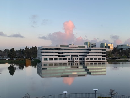 A modern office building reflected in a calm pond under a clear blue sky.