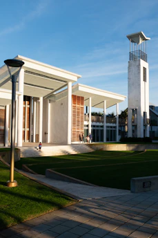 A modern architectural building with tall pillars and a large entrance is situated in an open area with neatly trimmed grass. A tall tower, resembling a clock tower, stands beside the building. A child walks on the steps leading to the building, and a few adults are seen near the entrance.