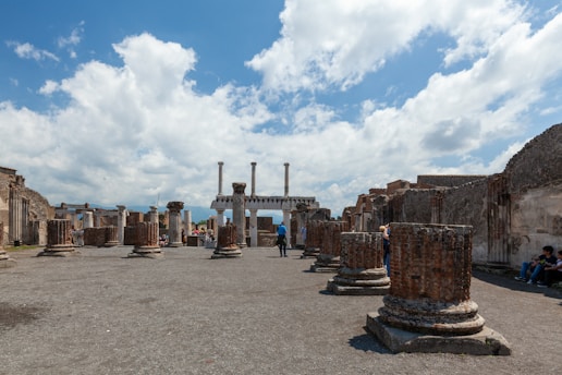 A historical archaeological site with ancient ruins, including stone columns and remnants of buildings under a partly cloudy blue sky. Visitors are scattered around, exploring the area.
