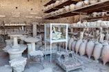 A room filled with ancient artifacts, including numerous amphorae and stone sculptures. These items are stacked on shelves and arranged on the floor. A glass case in the center displays a plaster cast of a figure, capturing a haunting pose. The stone walls in the background suggest the location is an archaeological site or a museum storage area.