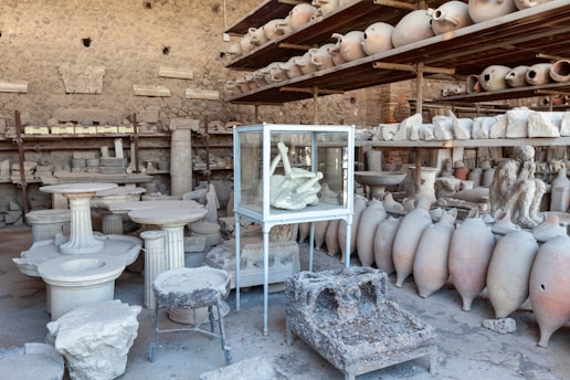A room filled with ancient artifacts, including numerous amphorae and stone sculptures. These items are stacked on shelves and arranged on the floor. A glass case in the center displays a plaster cast of a figure, capturing a haunting pose. The stone walls in the background suggest the location is an archaeological site or a museum storage area.