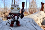 A toy figure with a detailed alien-like appearance, featuring a large head with yellow eyes and intricate armor, stands on a fallen log. The background consists of blurred trees and a clear sky, suggesting an outdoor setting.