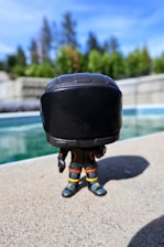 A toy figure with a large black helmet stands on a textured concrete surface, surrounded by greenery and a clear blue sky. The toy is accented with orange and yellow details on its suit.