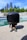 A toy figure with a large black helmet stands on a textured concrete surface, surrounded by greenery and a clear blue sky. The toy is accented with orange and yellow details on its suit.