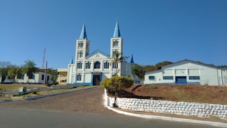 A large church with two tall spires and a light blue and white color scheme sits prominently on a hill. The surrounding area includes palm trees, a paved pathway leading up to the church, and adjacent buildings. The sky is clear and blue, adding to the serene and peaceful atmosphere.