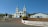 A large church with two tall spires and a light blue and white color scheme sits prominently on a hill. The surrounding area includes palm trees, a paved pathway leading up to the church, and adjacent buildings. The sky is clear and blue, adding to the serene and peaceful atmosphere.