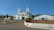 A large church with two tall spires and a light blue and white color scheme sits prominently on a hill. The surrounding area includes palm trees, a paved pathway leading up to the church, and adjacent buildings. The sky is clear and blue, adding to the serene and peaceful atmosphere.