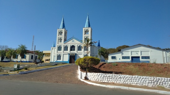 A large church with two tall spires and a light blue and white color scheme sits prominently on a hill. The surrounding area includes palm trees, a paved pathway leading up to the church, and adjacent buildings. The sky is clear and blue, adding to the serene and peaceful atmosphere.