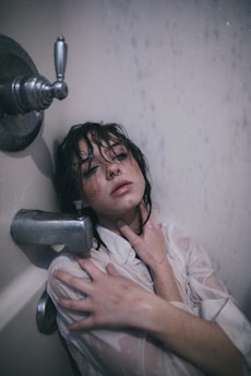 A person is sitting under a shower, fully clothed with wet hair and a white shirt. Their expression appears introspective or melancholic, with one hand on their chest and the other resting on their shoulder.