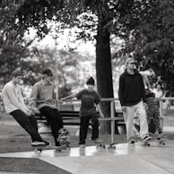 A group of five young individuals are engaging in a skateboarding session at a skatepark. They are casually dressed, wearing hats or beanies, and are positioned either sitting or standing on their skateboards. The background shows trees and a bench, creating a laid-back and leisurely atmosphere.