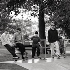 A group of five young individuals are engaging in a skateboarding session at a skatepark. They are casually dressed, wearing hats or beanies, and are positioned either sitting or standing on their skateboards. The background shows trees and a bench, creating a laid-back and leisurely atmosphere.