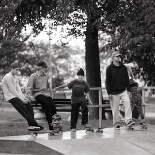 A group of five young individuals are engaging in a skateboarding session at a skatepark. They are casually dressed, wearing hats or beanies, and are positioned either sitting or standing on their skateboards. The background shows trees and a bench, creating a laid-back and leisurely atmosphere.
