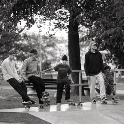 A group of five young individuals are engaging in a skateboarding session at a skatepark. They are casually dressed, wearing hats or beanies, and are positioned either sitting or standing on their skateboards. The background shows trees and a bench, creating a laid-back and leisurely atmosphere.