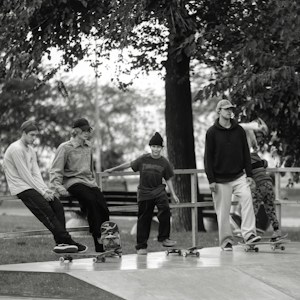 A group of five young individuals are engaging in a skateboarding session at a skatepark. They are casually dressed, wearing hats or beanies, and are positioned either sitting or standing on their skateboards. The background shows trees and a bench, creating a laid-back and leisurely atmosphere.