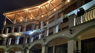 A multi-story building featuring an elegant design with arched balconies and stylish black wrought-iron railings. The top level is adorned with a wooden ceiling and warm string lights glowing softly, creating a cozy and inviting atmosphere. Potted plants are strategically placed on the balconies, adding a touch of greenery. The lighting on the facade creates a play of shadows, enhancing the architectural details.