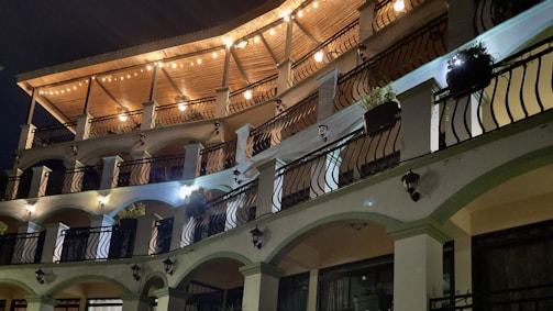 A multi-story building featuring an elegant design with arched balconies and stylish black wrought-iron railings. The top level is adorned with a wooden ceiling and warm string lights glowing softly, creating a cozy and inviting atmosphere. Potted plants are strategically placed on the balconies, adding a touch of greenery. The lighting on the facade creates a play of shadows, enhancing the architectural details.