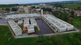 Aerial view of a residential complex with multiple identical buildings arranged in a geometric pattern. The structures are surrounded by paved areas and green spaces with dense vegetation beyond the walled perimeter. Adjacent to the complex are scattered houses and buildings in a suburban area.