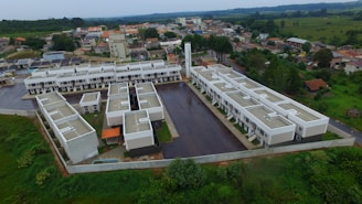 Aerial view of a residential complex with multiple identical buildings arranged in a geometric pattern. The structures are surrounded by paved areas and green spaces with dense vegetation beyond the walled perimeter. Adjacent to the complex are scattered houses and buildings in a suburban area.