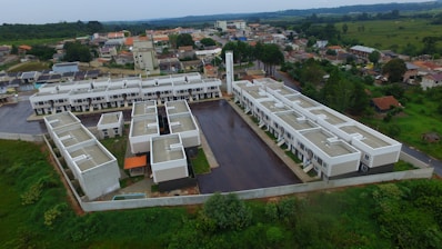 Aerial view of a residential complex with multiple identical buildings arranged in a geometric pattern. The structures are surrounded by paved areas and green spaces with dense vegetation beyond the walled perimeter. Adjacent to the complex are scattered houses and buildings in a suburban area.