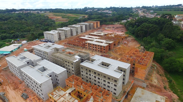 Multiple unfinished buildings are under construction, surrounded by forested and open land. The structures are at various stages, with some having complete frames and others still with exposed red brickwork.