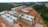 Multiple unfinished buildings are under construction, surrounded by forested and open land. The structures are at various stages, with some having complete frames and others still with exposed red brickwork.