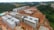 Multiple unfinished buildings are under construction, surrounded by forested and open land. The structures are at various stages, with some having complete frames and others still with exposed red brickwork.