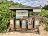 A set of five wooden recycling bins is situated outdoors on a sandy surface, surrounded by lush green foliage. Each bin is labeled for different types of waste: organic, cans, plastic, and glass. Above the bins, a wooden shelter provides protection, and there is informational signage with guidance on waste separation.