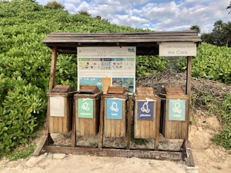 A set of five wooden recycling bins is situated outdoors on a sandy surface, surrounded by lush green foliage. Each bin is labeled for different types of waste: organic, cans, plastic, and glass. Above the bins, a wooden shelter provides protection, and there is informational signage with guidance on waste separation.