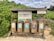 A set of five wooden recycling bins is situated outdoors on a sandy surface, surrounded by lush green foliage. Each bin is labeled for different types of waste: organic, cans, plastic, and glass. Above the bins, a wooden shelter provides protection, and there is informational signage with guidance on waste separation.