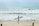 A person stands on a sandy beach holding a surfboard, gazing at the waves ahead. Other surfers can be seen in the water, riding the gentle waves under a cloudy sky.