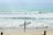 A person stands on a sandy beach holding a surfboard, gazing at the waves ahead. Other surfers can be seen in the water, riding the gentle waves under a cloudy sky.