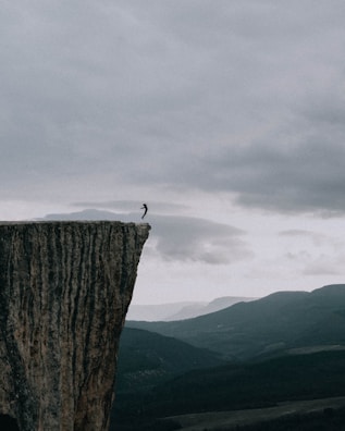 Wide-angle shot of a helicopter hovering above a cliff, with a jumper silhouetted against the sky.