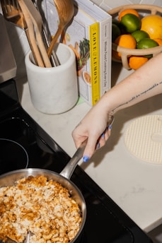 A person with blue nail polish holds a frying pan filled with macaroni and cheese with a crispy topping over a stove. Nearby, a marble container holds wooden utensils. A cookbook is propped open, and a basket with oranges and limes sits on the counter. A tattoo is visible on the person's arm.