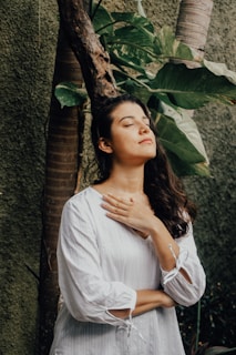 A person with long dark hair, wearing a white blouse, stands with eyes closed and hands on their chest. The background includes green foliage and tree trunks, creating a natural ambiance.