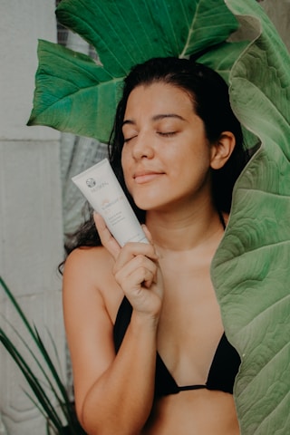 A person in a black bikini holds a skincare product near their face, surrounded by large green leaves. Their eyes are closed, and they appear relaxed and content.