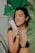 A person in a black bikini holds a skincare product near their face, surrounded by large green leaves. Their eyes are closed, and they appear relaxed and content.